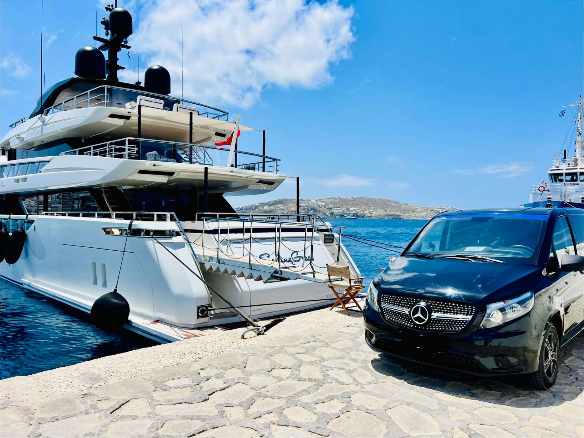 Luxury white van on a Mykonos coastal road with white Cycladic buildings and blue sea